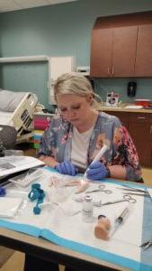 A person with gloves appears to be performing a medical procedure on a simulation model in a clinical setting, with various medical tools and equipment around them.